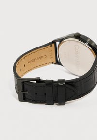 Μαύρο δερμάτινο λουρί ρολογιού Calvin Klein με μεταλλικό αγκράφα και ραφές, με το καντράν του ρολογιού να είναι γυρισμένο προς τα πίσω, αποκαλύπτοντας το όνομα της μάρκας.
