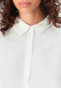 Camicia bianca con colletto classico, caratterizzata da una chiusura a bottoni frontale, tessuto morbido e un design semplice con bordi arrotondati del colletto e piccoli bottoni.