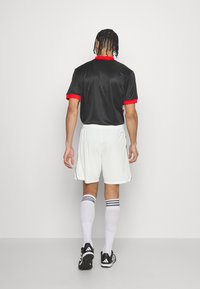 Jersey negro de manga corta con detalles en rojo, pantalones cortos blancos y zapatillas deportivas negras. Calcetines blancos con rayas negras y azules. Fondo simple.