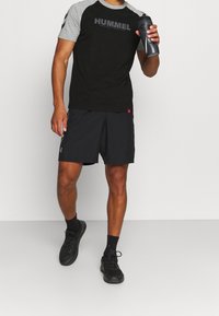 Sort og grå sportst-shirt med "HUMMEL" logo, parret med sorte shorts. Modellen holder en sort vandflaske og har sorte træningssko på.