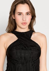 Top halter preto com design pregueado, apresentando um decote curvo e alças cruzadas. Textura suave e silhueta ajustada.