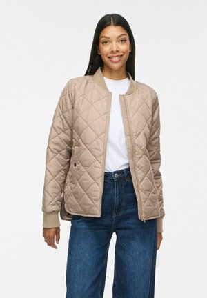 Femme souriante portant une veste matelassée beige sur une chemise blanche et un jean bleu, debout devant un fond clair uni.
