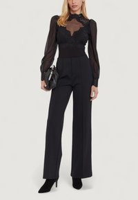 Kobieta w przezroczystej czarnej koronkowej bluzce z długim rękawem, w czarnych spodniaczh w stylu wide-leg z wysokim stanem, w czarnych botkach na obcasie i trzymająca czarną kopertówkę.