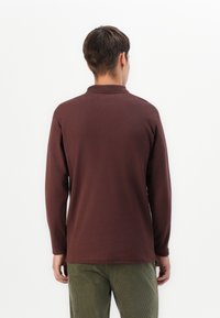 Cămașă polo cu mâneci lungi, de culoare maro închis, cu un material texturat, având un guler standard și o margine dreaptă, purtată cu pantaloni din corduroy de culoare măslin.