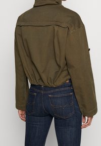 Olivgrön kort jacka med hög krage, elastisk nederkant och strukturerade axlar, kombinerad med mörka denimjeans med bakfickor.