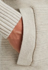 Beige gebreide trui met ribbelmanchetten en een zijzak. De gestructureerde stof heeft een zachte, geweven uitstraling met een comfortabele feel.