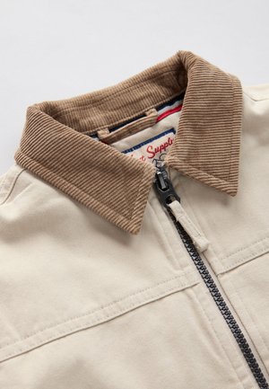 Bēša jaka ar brūnu korduroja apkakli. Tajā ir melna rāvējslēdzēja aizdare, divas priekšējās kabatas un redzama izšūšana uz auduma.