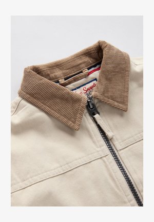 Bež jakna z rjavim velur ovratnikom. Ima črni zadrgo, dva sprednja žepa in vidno šivanje na tkanini.