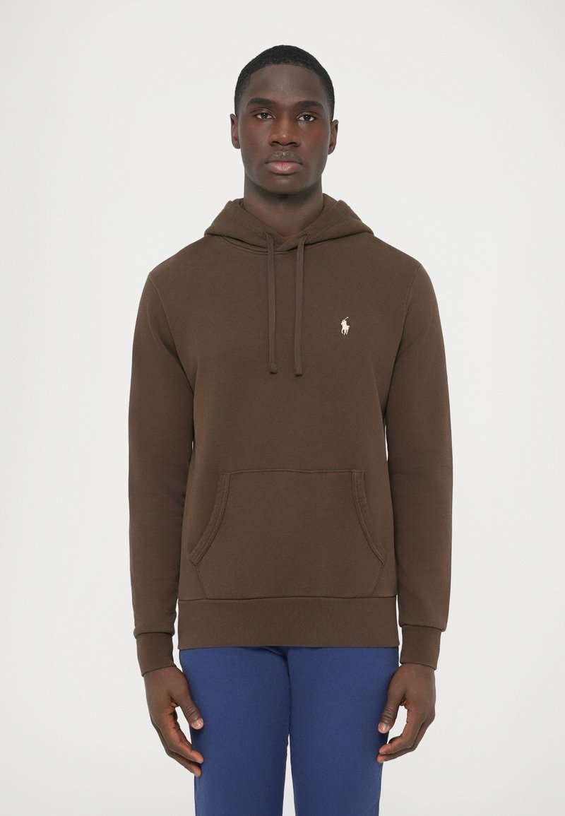 Barna pamut kapucnis pulóver húzózsinóros kapucnival, elülső zsebbel és bordázott mandzsettával. Hímzett logó a mellkason. Kék nadrággal viselve.