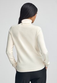 Donna con lunghi capelli scuri, che indossa un maglione a collo alto di colore crema e pantaloni neri, girata di spalle su uno sfondo grigio chiaro uniforme.
