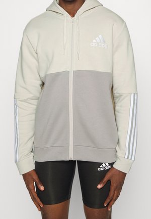 Man draagt een tweekleurige beige en grijze Adidas zip-up hoodie met drie strepen op de mouwen en zwarte Adidas shorts.