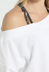 Bílý svetřík bez ramínek s texturovanou tkaninou, volným střihem a černými vzorovanými popruhy viditelnými na rameni.