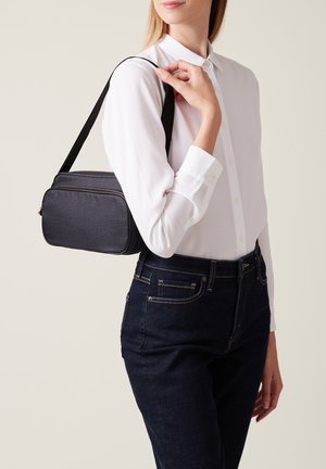 Borsa a tracolla in pelle nera con finitura testurizzata, forma rettangolare, chiusura con zip e tracolla regolabile, indossata sopra una camicia bianca e jeans.