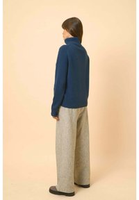 Tmavě modrý pletený rolák s žebrovanými manžetami, spárovaný se šedými širokými kalhotami a černou obuví, pohled zezadu.