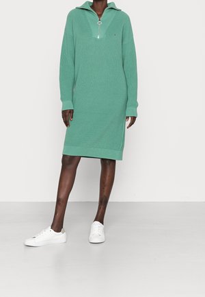 Hellgrünes Strickkleid mit halbem Reißverschluss am Kragen, geripptem Muster, langen Ärmeln und asymmetrischem Saum, kombiniert mit weißen Turnschuhen.