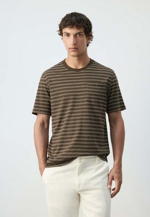Hombre con una camiseta de manga corta de rayas marrones, parcialmente metida en pantalones color crema, sobre un fondo claro y liso.