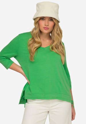 Groene langemouwen top van lichtgewicht materiaal met een V-hals, zijsplitten en een losse pasvorm. Gecombineerd met een witte bucket hat.
