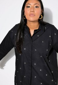 Camisa negra con ojales metálicos en un patrón disperso, diseño con cuello y mangas largas; textura suave con un ajuste estructurado.
