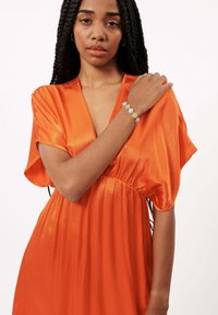 Oranges Satin-Kleid mit kurzen Ärmeln und taillierter Taille. Armband mit bunten Steinen in Grün, Blau und transparenten Farbtönen am Handgelenk.