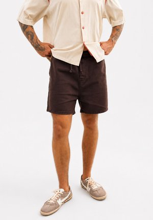 Homme portant une chemise beige à manches courtes boutonnée, un short marron foncé, des baskets beiges, debout les mains dans les poches sur fond blanc.