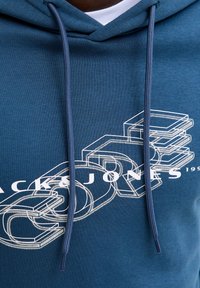 Μπλε φούτερ με κουκούλα που διαθέτει λευκό γραφικό σχέδιο με στοίβες γράμματα, κορδόνια και υφή υφάσματος. Το όνομα της μάρκας εμφανίζεται σε εξέχουσα θέση.