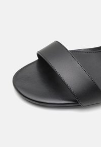 Gros plan sur une sandale à bout ouvert en cuir noir avec une large bride unique à l'avant et une semelle intérieure à texture lisse.