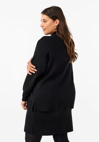 Schwarzer, übergroßer Pullover mit einem strukturierten, gerippten Saum, langen Ärmeln und einem lockeren Schnitt. Kombiniert mit einem passenden schwarzen Rock.