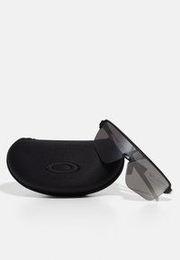 Sorte solbriller med et elegant, kantet design og grå linser, som hviler delvis inne i et svart mykt etui med en logo.