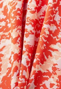 Tissu aux plis drapés présentant un motif camouflage abstrait dans des nuances de rouge, orange et rose clair.
