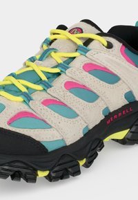 Chaussure de trail avec tige beige, sarcelle et rose, lacets jaune vif et semelle noire robuste avec accents jaunes.