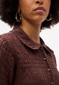 Καφέ πλεκτό πουλόβερ με γιακά και λεπτομέρεια κουμπιού. Μοντέλο με αφηρημένα χρυσά σκουλαρίκια σε σχήμα κρίκου. Κοντινή λήψη.