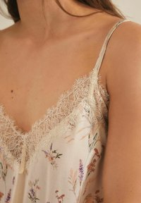 Camisole floral en tissu crème clair, avec une finition en dentelle le long du décolleté. Motif floral délicat dans des couleurs pastel douces. Bretelles ajustables.