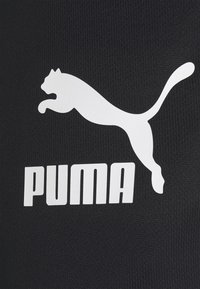 Černé sportovní oblečení s bílým logem Puma, na kterém je nad názvem značky v tučném fontu grafika skákající pumy. Texturovaný povrch látky.