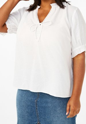 Vrouw die een wit blouse met korte mouwen en een gestrikte halsband draagt, samen met een blauwe spijkerrok, poserend met één arm omhoog.