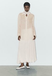 Helle beige, durchsichtige Langarmbluse mit Kragen, kombiniert mit einem fließenden, knöchellangen Rock in passender Farbe; flache, cremefarbene Schuhe runden den Look ab.