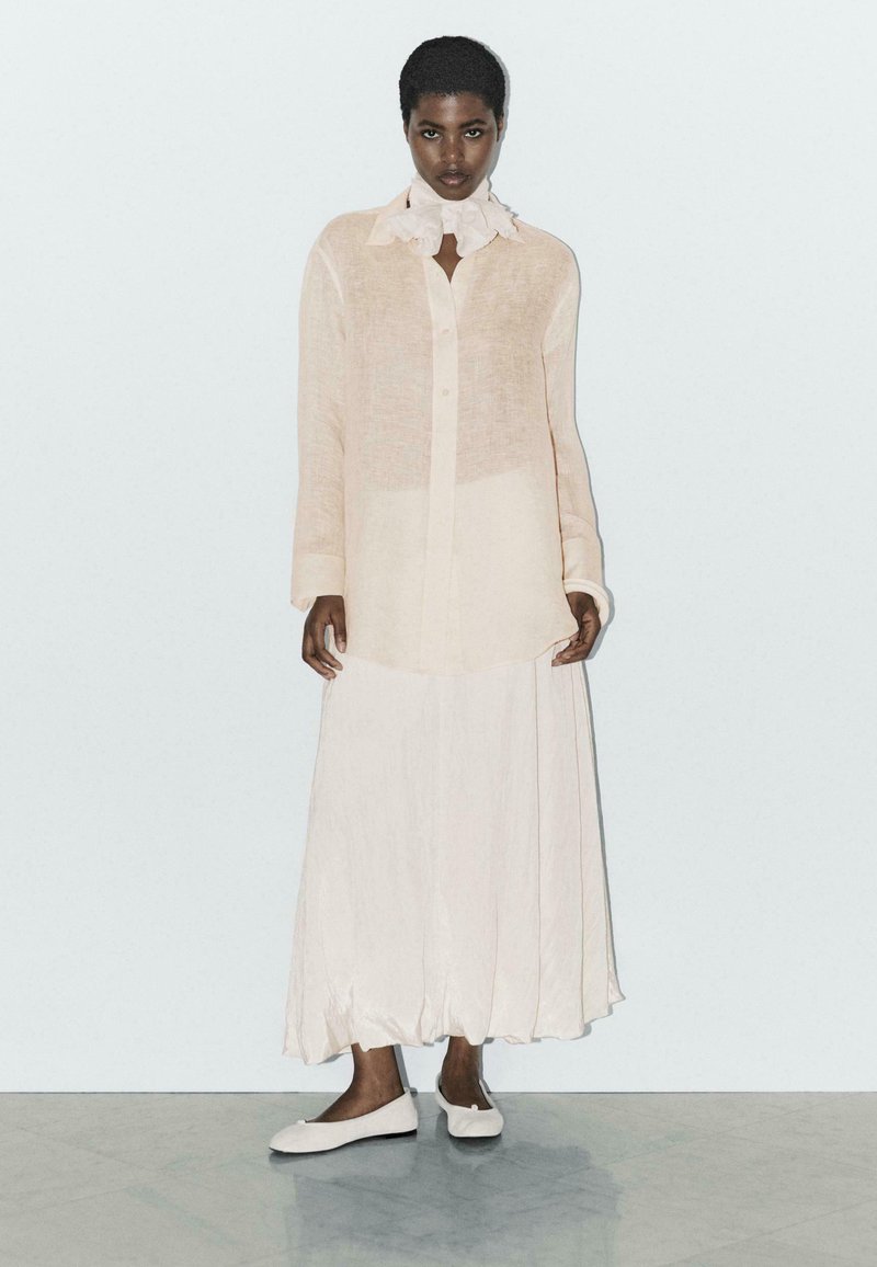 Helle beige, durchsichtige Langarmbluse mit Kragen, kombiniert mit einem fließenden, knöchellangen Rock in passender Farbe; flache, cremefarbene Schuhe runden den Look ab.