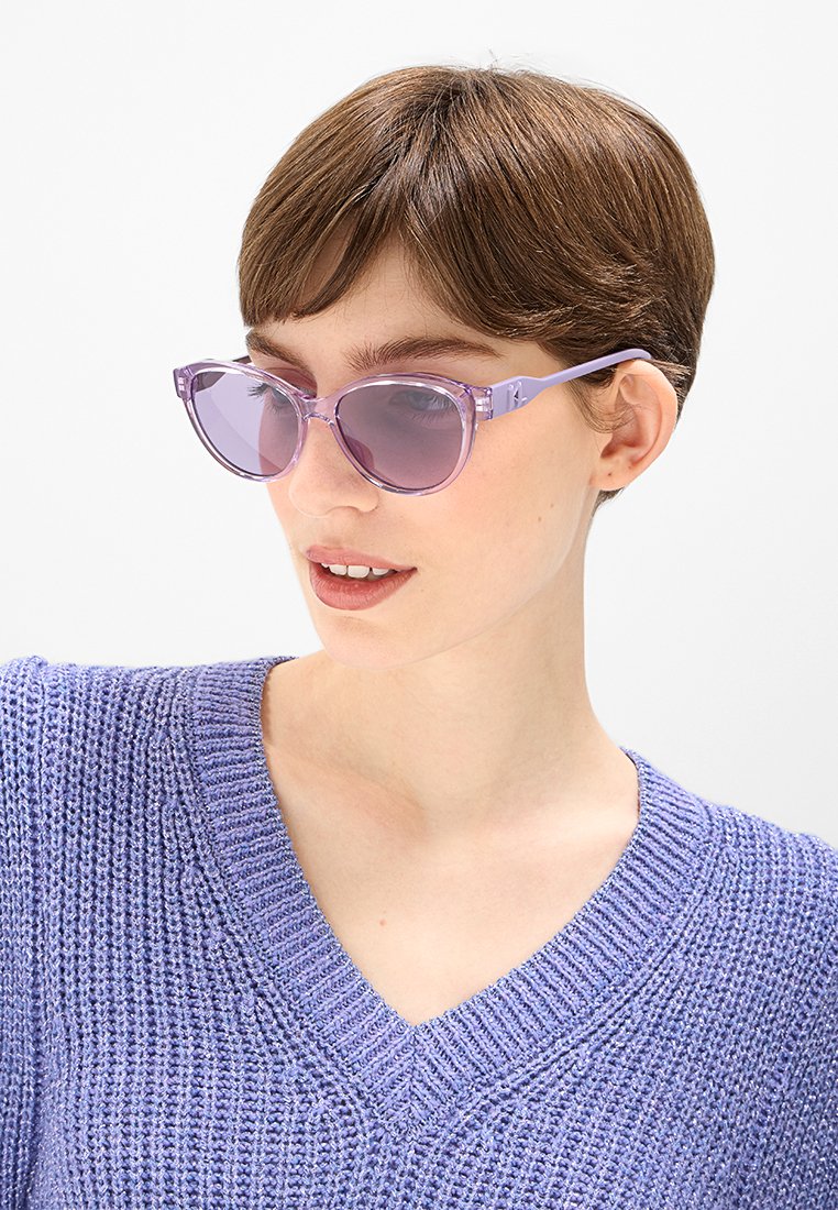 Lunettes de soleil en forme de yeux de chat, translucides et violettes, avec des verres dégradés et des montures en plastique légères. Portées avec un pull texturé lavande.