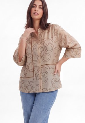 Vrouw draagt een beige blouse met knopen en swirlpatronen en blauwe spijkerbroek, poseert met één hand op de heup en de andere bij de kraag.