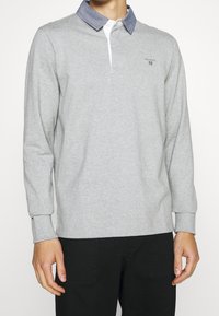 Vaaleanharmaa pitkähihainen polo-paita, jossa on tummansinisestä chambray-kankaasta valmistettu kaulus ja valkoinen napitus. Hartialla on kirjontaa.