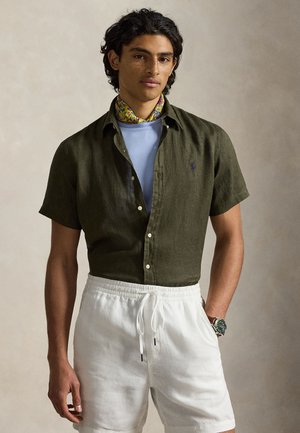 Młody mężczyzna ubrany w oliwkową, krótka koszulę na guziki, niebieską podkoszulkę, białe szorty z troczkami, kwiatowy szalik na szyi i zegarek na nadgarstku, stoi w pomieszczeniu.