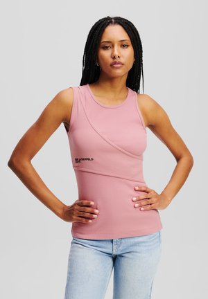 Roze mouwloos topje met een geribbelde structuur, voorzien van een asymmetrisch ontwerp en een subtiele logo-detail aan de voorkant. Gemaakt in combinatie met lichtblauwe spijkerbroek.