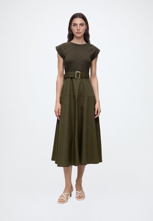 Femme aux longs cheveux foncés porte une robe midi sans manches vert olive avec une large ceinture et des sandales beiges à talons et à brides, debout sur un fond blanc.