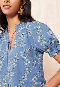 Blusa azzurro chiaro con motivo floreale bianco e giallo, maniche puff corte e scollatura a V. Tessuto liscio con un leggero luccichio.