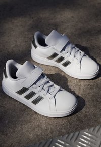 Bijele sportske tenisice s glatkim kožnim gornjim dijelom, mrežastim jezikom, tri metalne srebrne pruge i gumeni potplat. Imaju vezice i čičak traku.