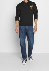 Felpa nera con cappuccio e tasca frontale, caratterizzata da tre strisce a chevron in grigio e arancione. Abbinata a jeans blu e sneakers nere.