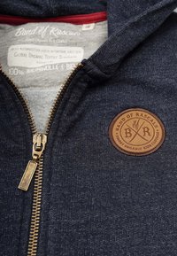 Marineblå zip-up hoodie lavet af 100% økologisk bomuld, med et struktureret stof, bronze lynlås og et rundt brunt logo-patch.