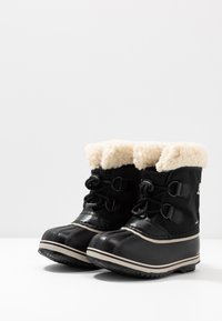 Svarte vanntette snøboots med hvit syntetisk pelsfor, teksturerte gummisåler og snøring; distinkt blankt overmateriale.