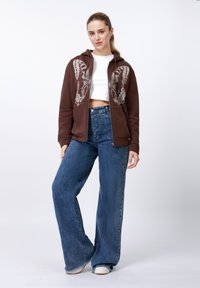 Brūnas zip-uzvilkamas kapuces džemperis ar tauriņa grafiku, kopā ar augsta vidukļa zilām plašām džinsām un baltām slip-on kurpēm.