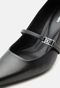 Μαύρο δερμάτινο παπούτσι με μύτη, επίπεδο με λεπτό λουράκι και ασημένια πόρπη στην κορυφή.