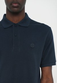 Mørkeblå polo skjorte med teksturert stoff, flat krage og tre knapper foran. Har en tonet logo på venstre bryst.